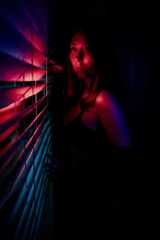 a girl poses with her hand on the blinds in a dark room