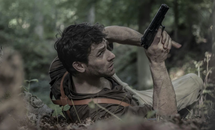 a young man sitting on a leaf covered field holding a gun