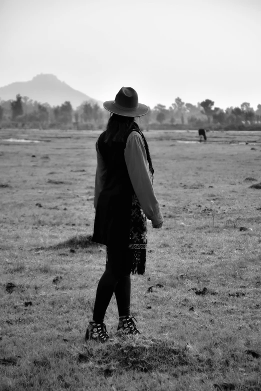 a black and white po of a woman wearing a cowboy hat and coat looking at a cow grazing in the distance