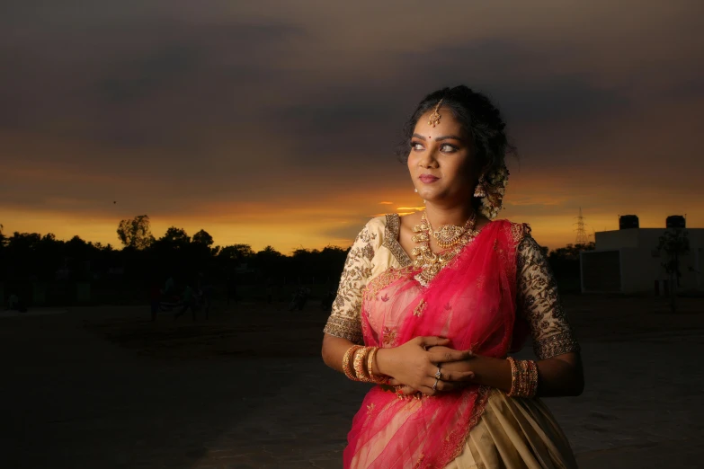 a woman wearing a pink and beige sari standing outside with her hands folded on the chest