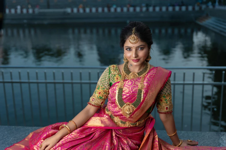 an indian woman wearing a red and gold saree sitting in front of a lake