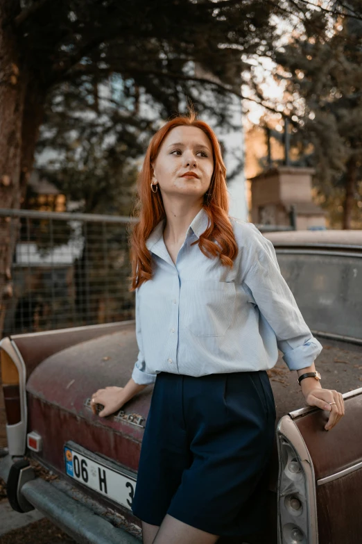 a red - headed woman leans on the front hood of a rusty car