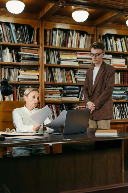 a man and woman looking at papers near a laptop on a desk in a liry