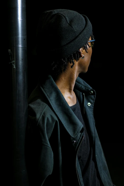 a woman with a jacket and hat looks ahead while standing in the dark