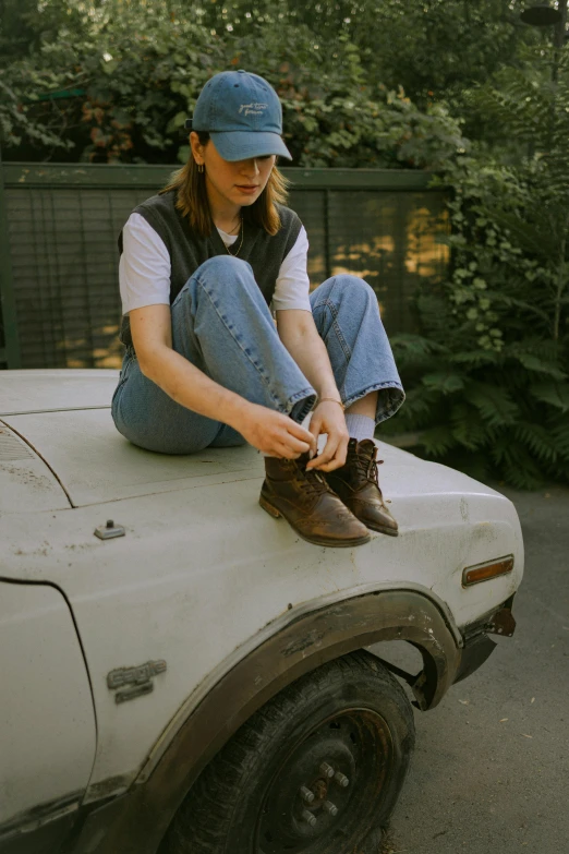 a woman sits on the bonnet of an old car, and fixes her shoelaces