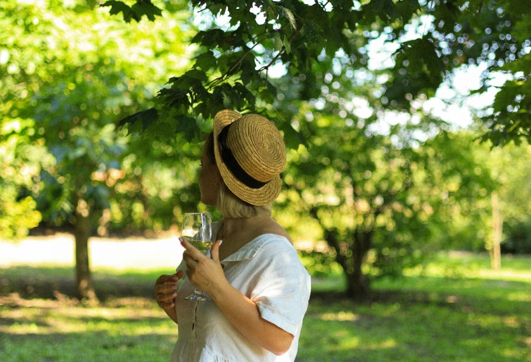 a woman wearing a hat standing in front of a tree holding a glass