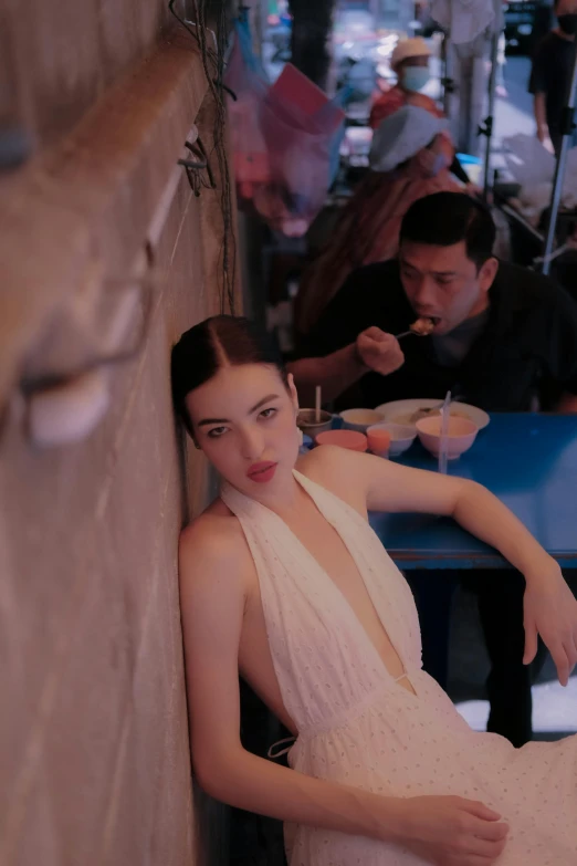 a woman leaning against a wall and eating food with a man at a table behind her