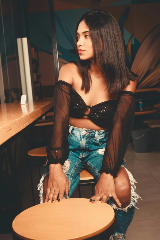 a woman in a blouse and jean shorts sits on a stool at a table