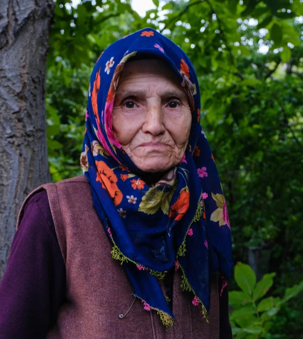 an elderly woman with a scarf on stands next to a tree