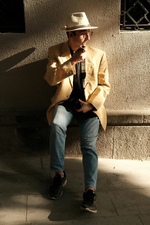 a man with glasses sits on a bench, smoking a cigarette and wearing a hat