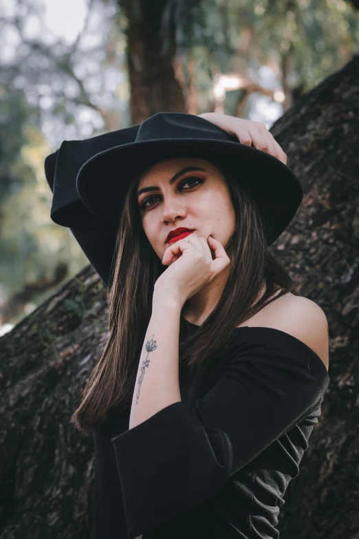 a woman in a black dress and hat poses near a tree