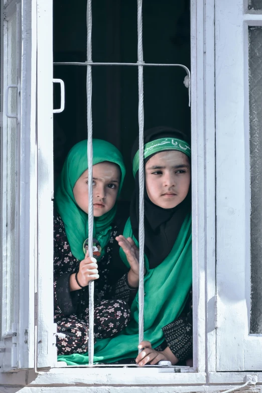 two girls in green headscarves look out a window