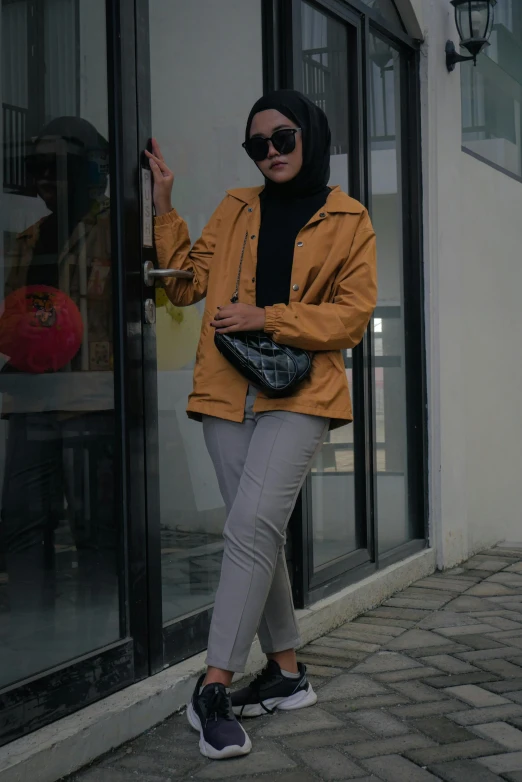 a woman in a hijab and sunglasses holding a purse is posing in front of glass doors