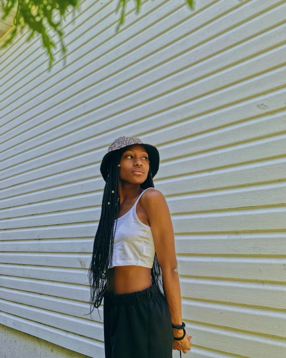 a woman standing in front of a white wall wearing black pants and a hat