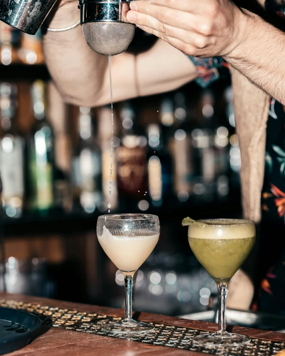 two different colored cocktails sit on a bar, one being poured from the cup, and the other is being filled