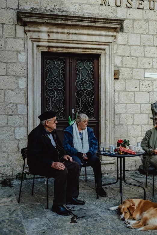 several older people sit outside a building while a dog lays on the ground