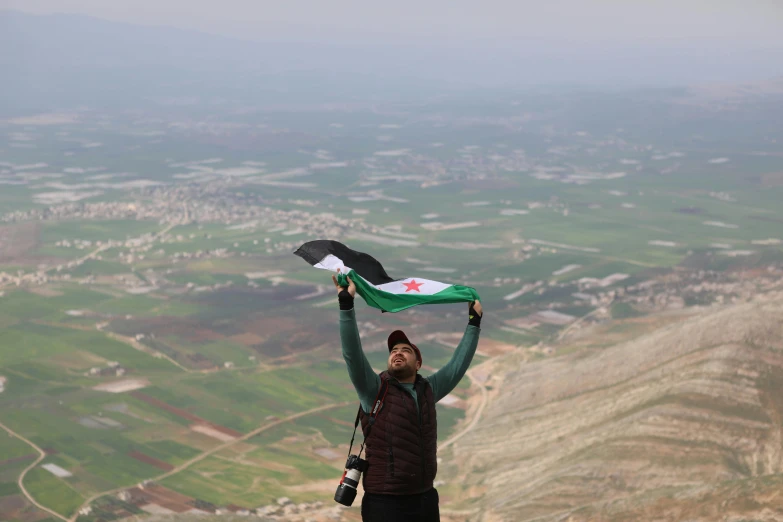 a man with an afghan flag holds his hands up above a valley and town