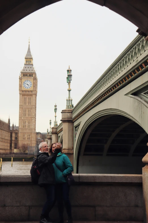 two people standing on the side walk of an arch next to a clock tower