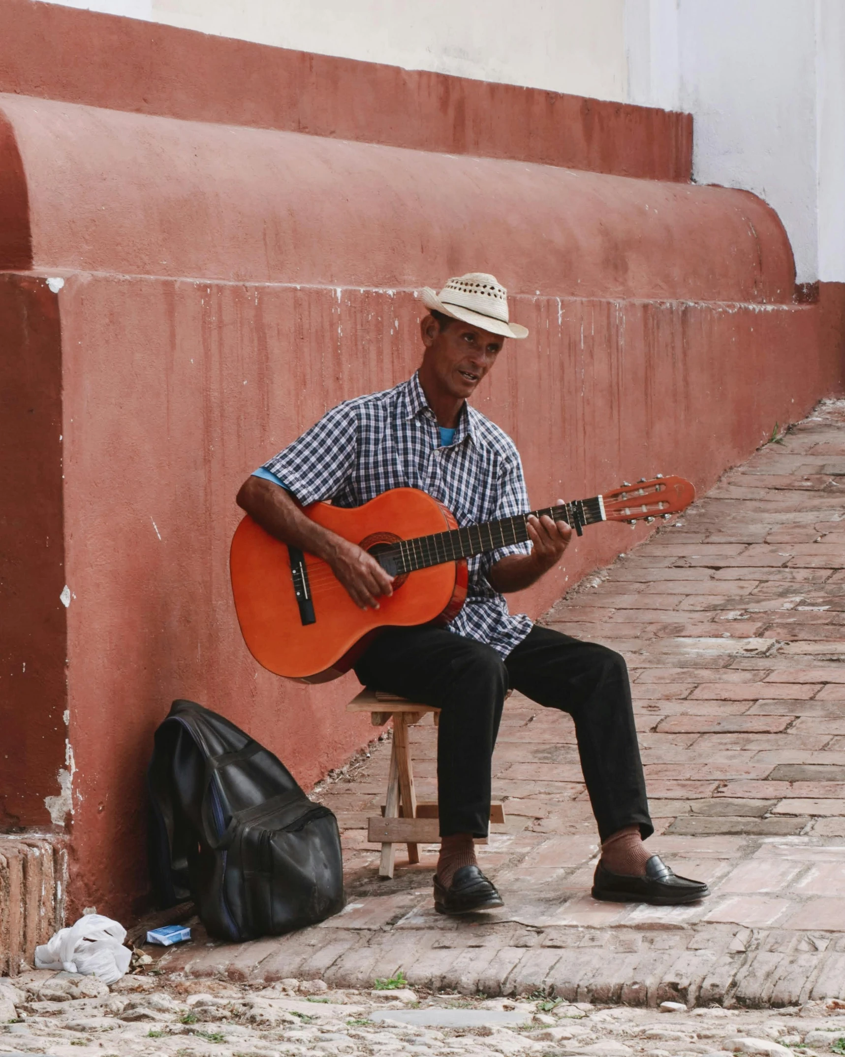 a man in hat sitting with guitar next to cement wall