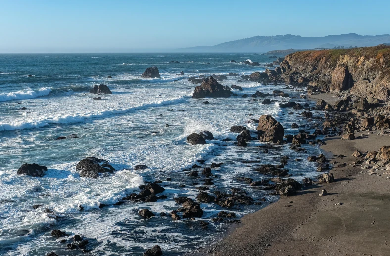 the coast with blue water has rocks on the shore and hills in the background