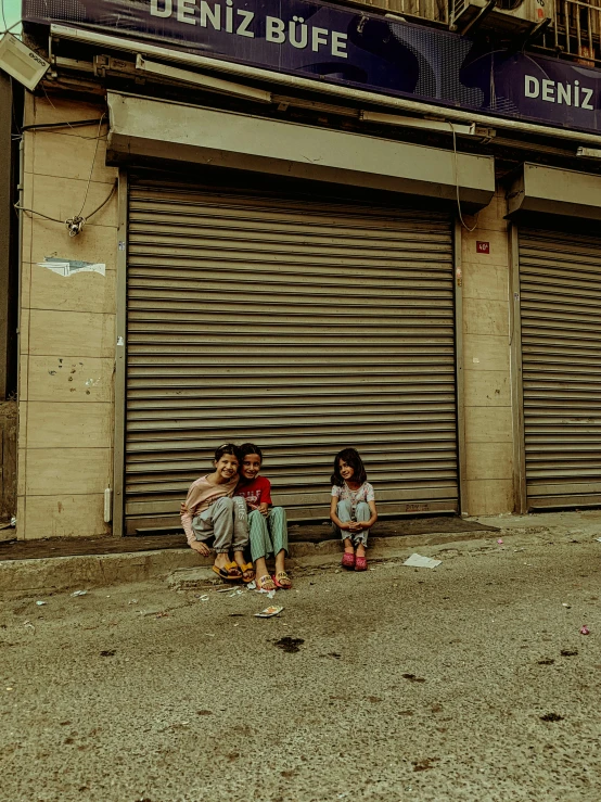 two girls sit on the sidewalk outside a closed shop