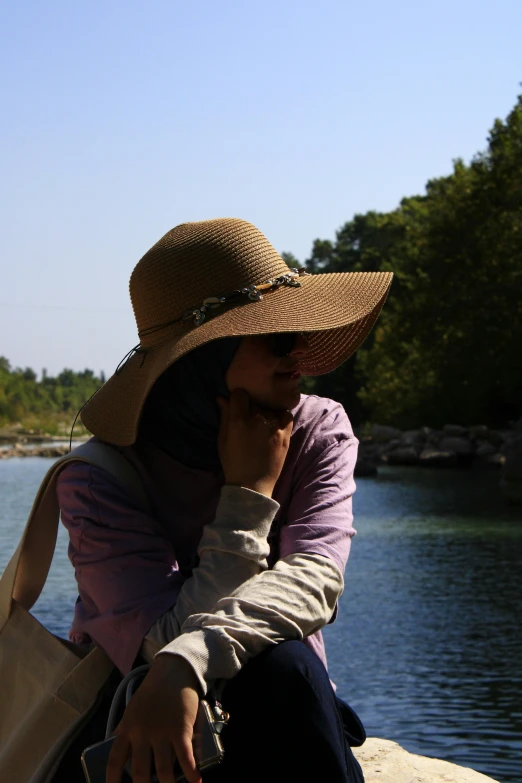 woman in brown hat sitting by water with cell phone