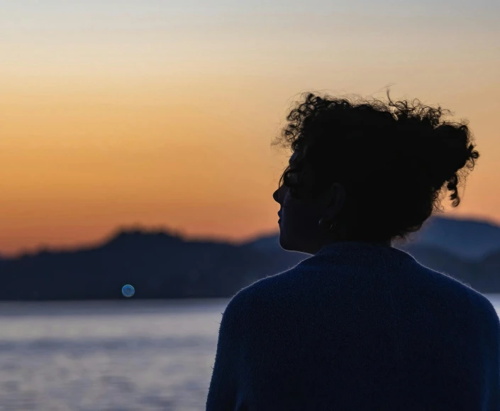 a woman with curly hair is silhouetted against the sun setting over a lake