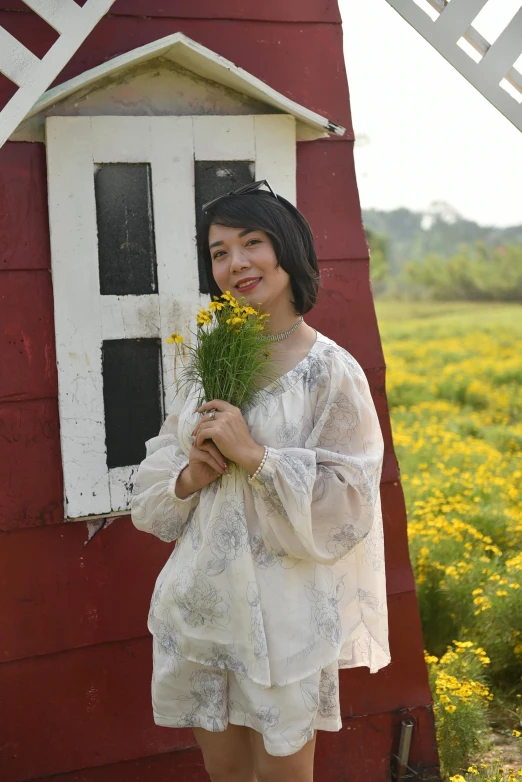 a beautiful woman in white standing next to a barn holding yellow flowers