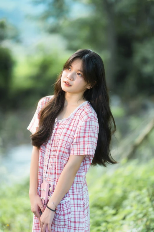 an asian girl stands by the grass and trees with her long hair in a ponytail