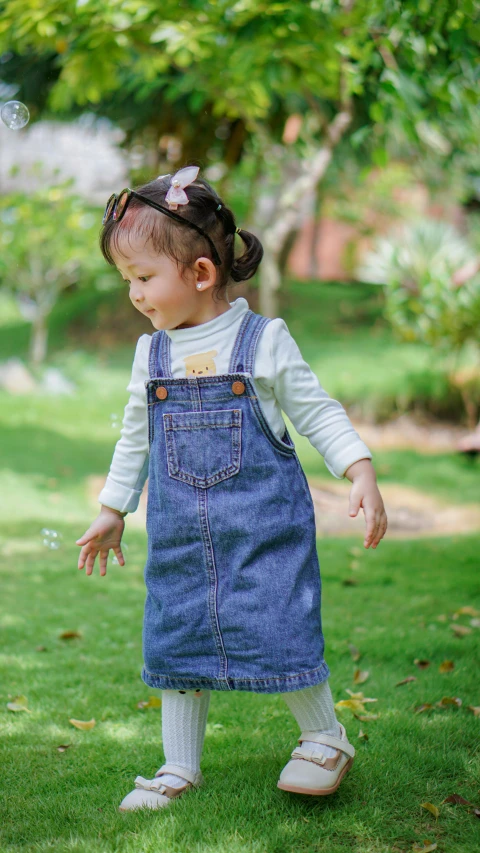 a  in a blue jean overall dress, with her arms extended and her leg up against the grass