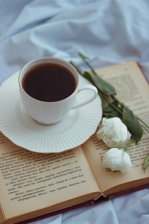 an open book is on the edge of the table with a cup of coffee and white flowers