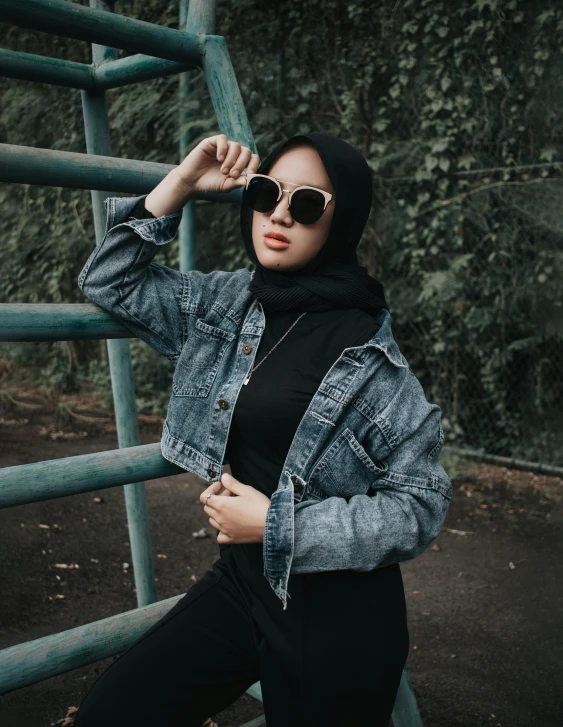 a woman in sunglasses and a denim jacket poses by a metal gate