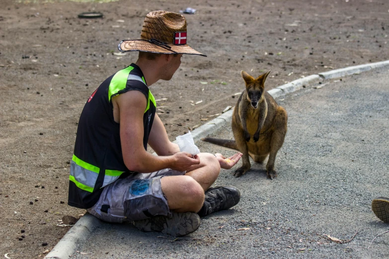 a man in a hat sitting on the side of a road next to a kangaroo