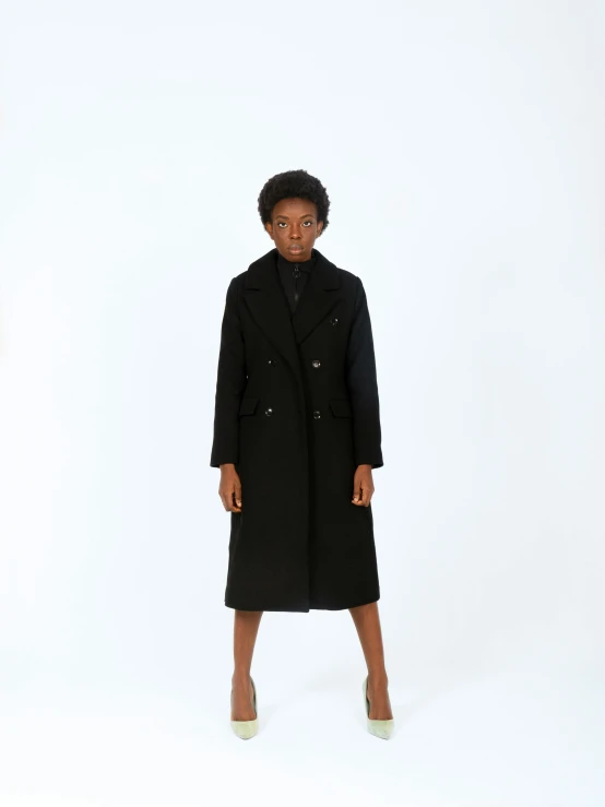 a woman wearing a black coat and standing in front of a white wall