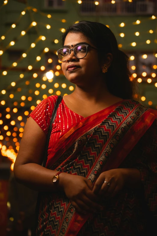 a woman wearing a red and brown blouse, with eye glasses standing in front of fairy lights