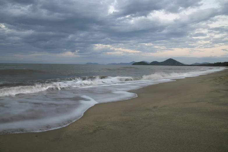 a sandy beach with a wave coming in and some mountain peaks visible