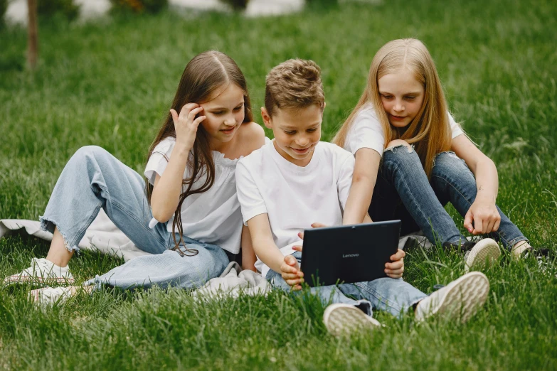 three children sitting on the grass looking at a laptop