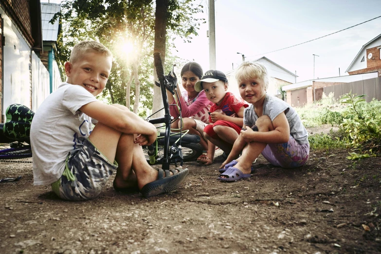 three children sitting on a dirt road with one girl on the ground and two boys on the front