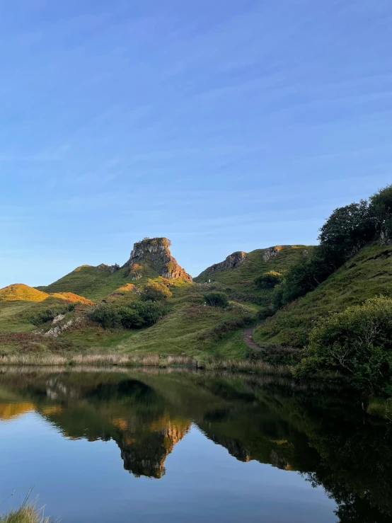 a pond on the side of a green hill with a tower that stands on a small island