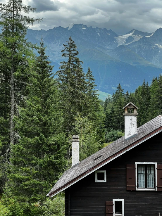 a cabin at the bottom of a mountain with tall trees and snow capped mountains in the background