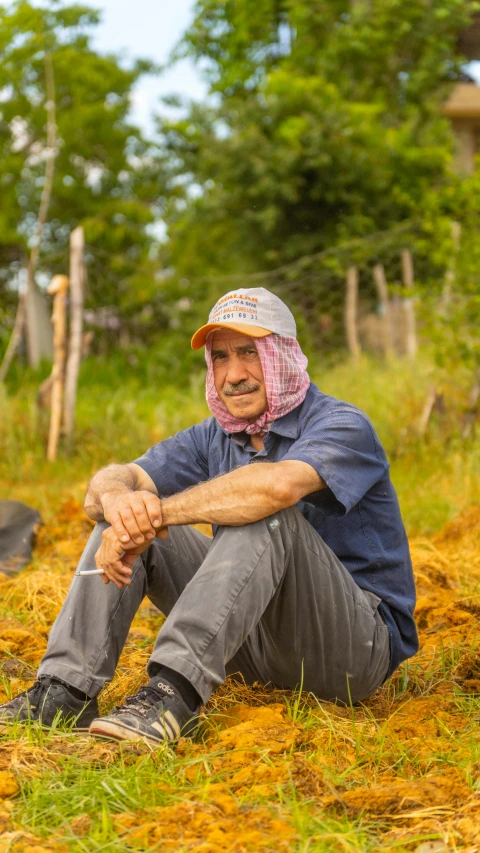 man with pink scarf and hat sitting in the grass on a farm