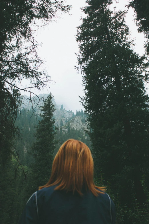 a girl stands in front of some trees looking over the hills