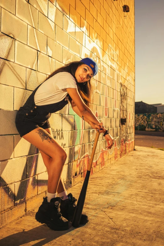 a woman in short shorts and baseball cap standing next to a brick wall with a bat