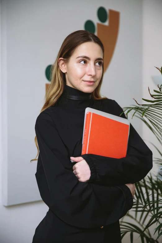 a woman holds a red notebook while standing next to a plant