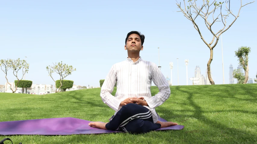 a man in white shirt and blue pants sitting on yoga mat with trees in background