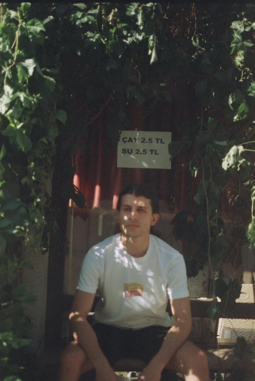 a man sitting down by a bush with a sign on his head