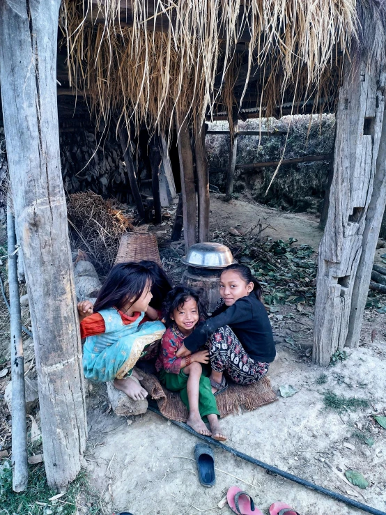 three children sitting under a shelter in a dirt area