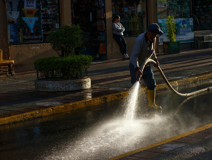 a man in shorts and an open cap hoses water into a street