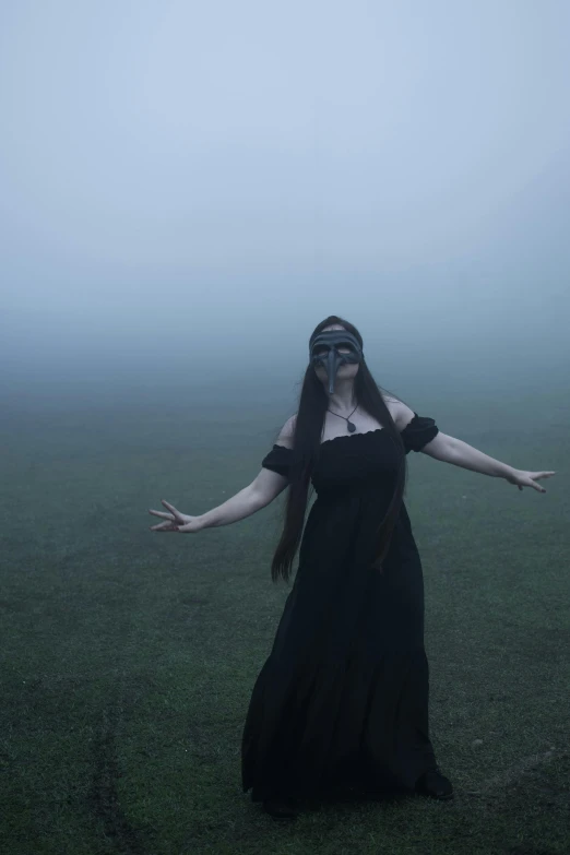a woman in black dress and blindfolded head standing on grassy field