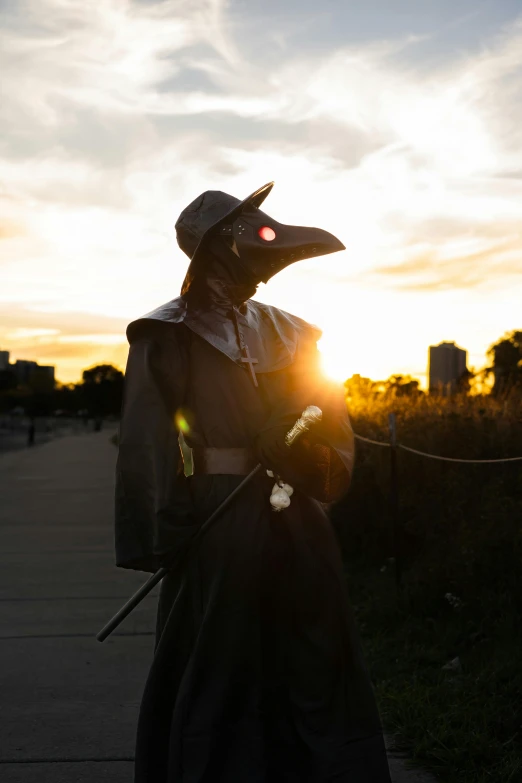 a person in a hooded outfit stands on a sidewalk with the sun behind them